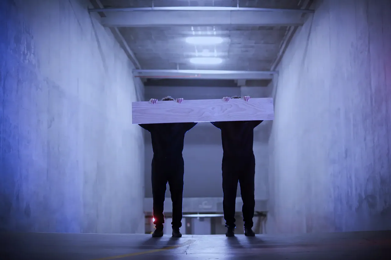 Two figures carry a board in a parking garage, Los Innato series